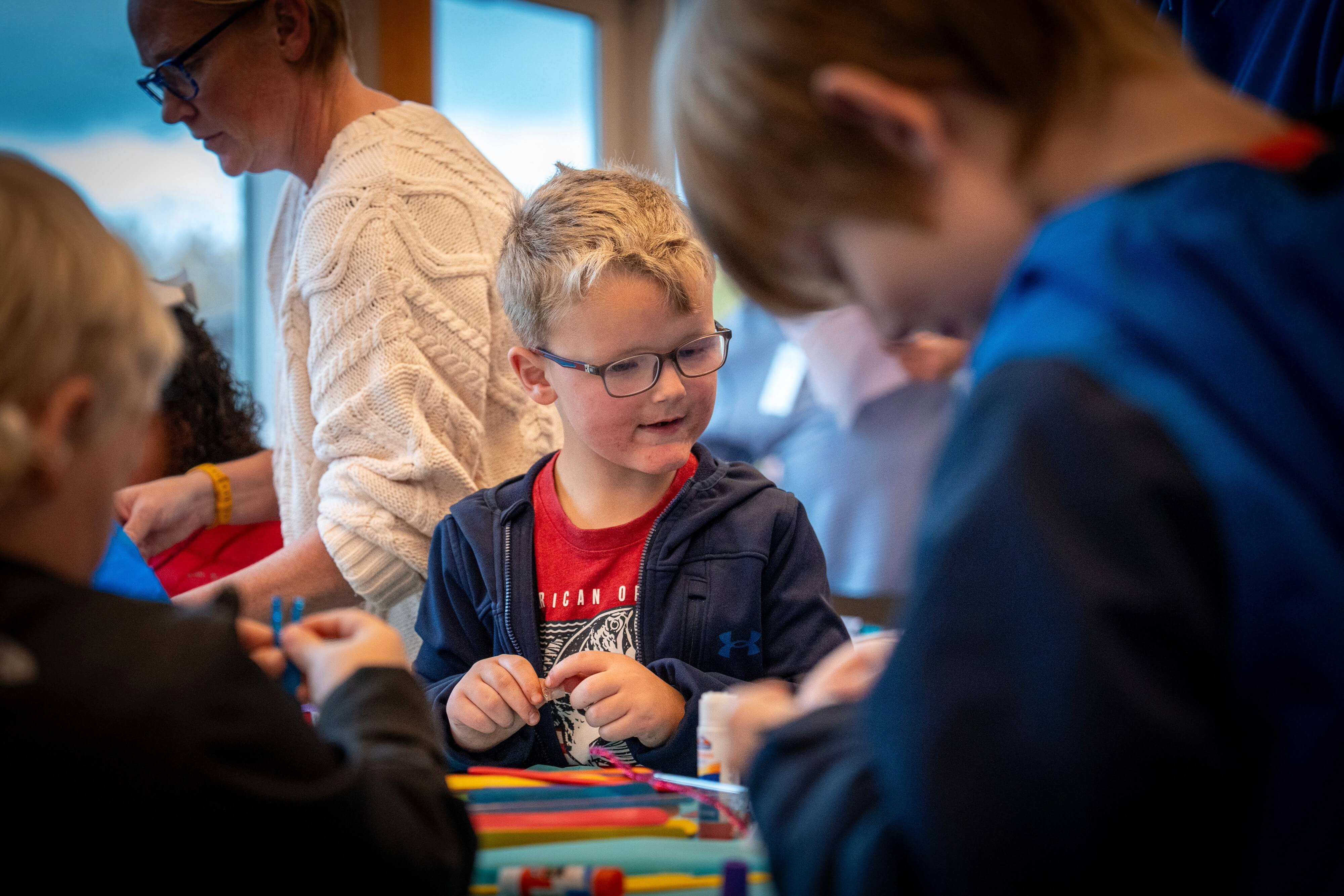 A young boy wearing glasses works on a craft project at a table filled with colorful supplies, surrounded by other children and adults.