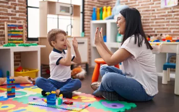 Teacher and toddler sitting on floor high five with hands raised up at kindergarten.