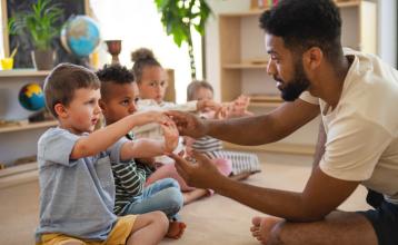 Group of small nursery school children with male teacher sitting on floor doing exercise.