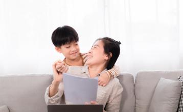 A mom sitting on sofa with her son while she is typing on laptop.