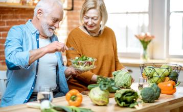 A senior couple eating salad standing together with healthy food on the kitchen at home.