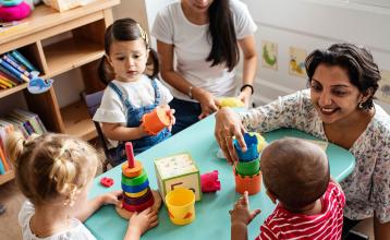 Teacher stacks cups with a child at a table while two other children play with toys