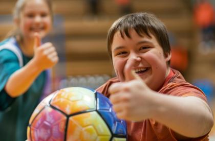 two kids one in the front holding a soccer ball giving a thumbs up. The other kid is in the background giving a thumbs up too. 
