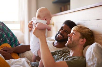 Two dads in bed, holding and smiling at their baby