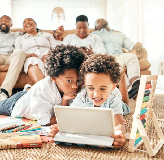Two children playing on laptop with family members on couch behind them.