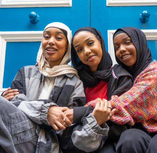 Three smiling women wearing hijabs embracing in city.
