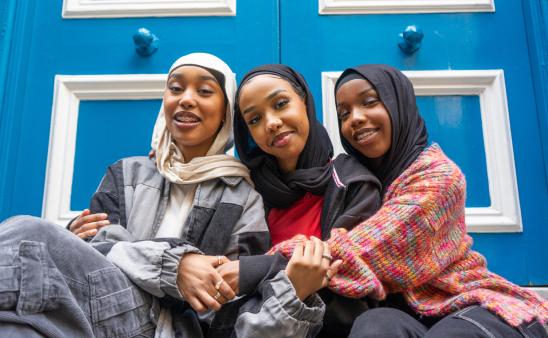 Three smiling women wearing hijabs embracing in city.