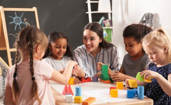 A child care provider sitting at a table with a group of children playing with blocks.