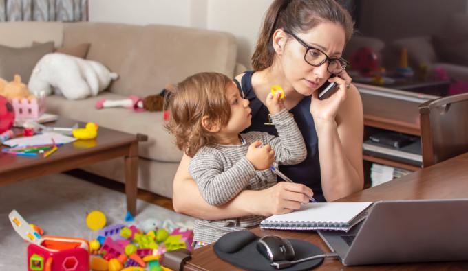 Young woman talking on phone and working on a laptop with child in her lap.