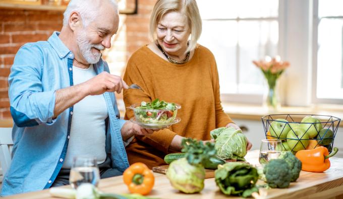 A senior couple eating salad standing together with healthy food on the kitchen at home. 