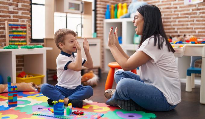 Teacher and toddler sitting on floor high five with hands raised up at kindergarten.