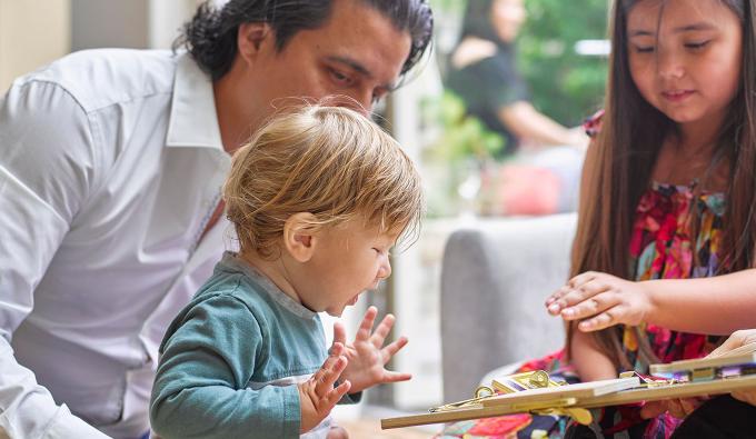 Toddler excited by a toy an older child is holding while a parent looks on