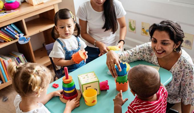 Teacher stacks cups with a child at a table while two other children play with toys 