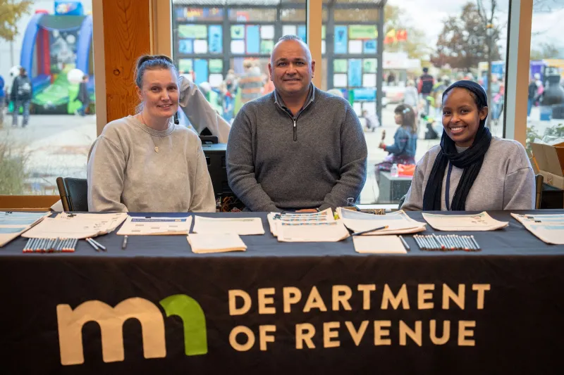 Three Minnesota Department of Revenue staff sit behind an information table covered with brochures and forms. The tablecloth displays the department’s logo, and event activities are visible through the windows behind them.