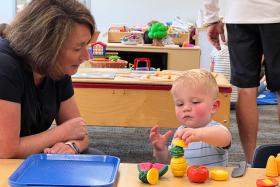 Toddler playing with wooden fruits while a caregiver watches