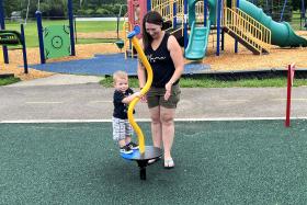 Mom stands close by making sure toddler doesn't fall while standing on a circular platform with a yellow pole