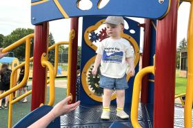 Toddler wearing hat stands on the jungle gym while a parent's arm reaches out to help
